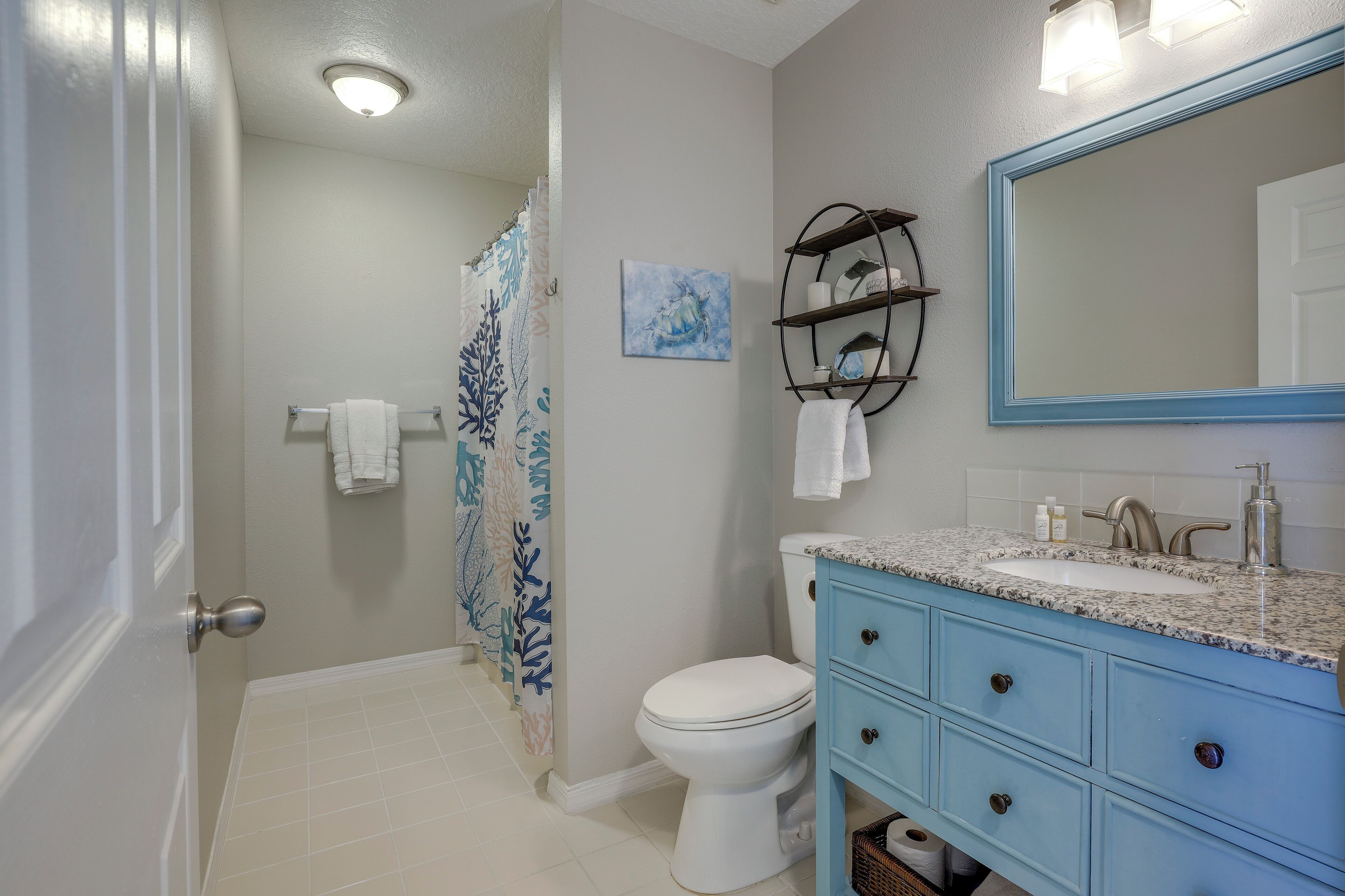 A bathroom with a white toilet, a blue vanity with a sink, a mirror, and a shower curtain with a leaf pattern.