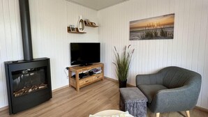 Una sala de estar con un estufa de leña negra, un televisor de pantalla plana sobre un soporte de madera, un sillón gris y un cuadro de un atardecer en la pared.