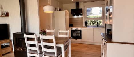 Una cocina moderna con armarios blancos, horno empotrado y estufa negra. Hay una mesa de comedor con sillas blancas y una lámpara colgante.