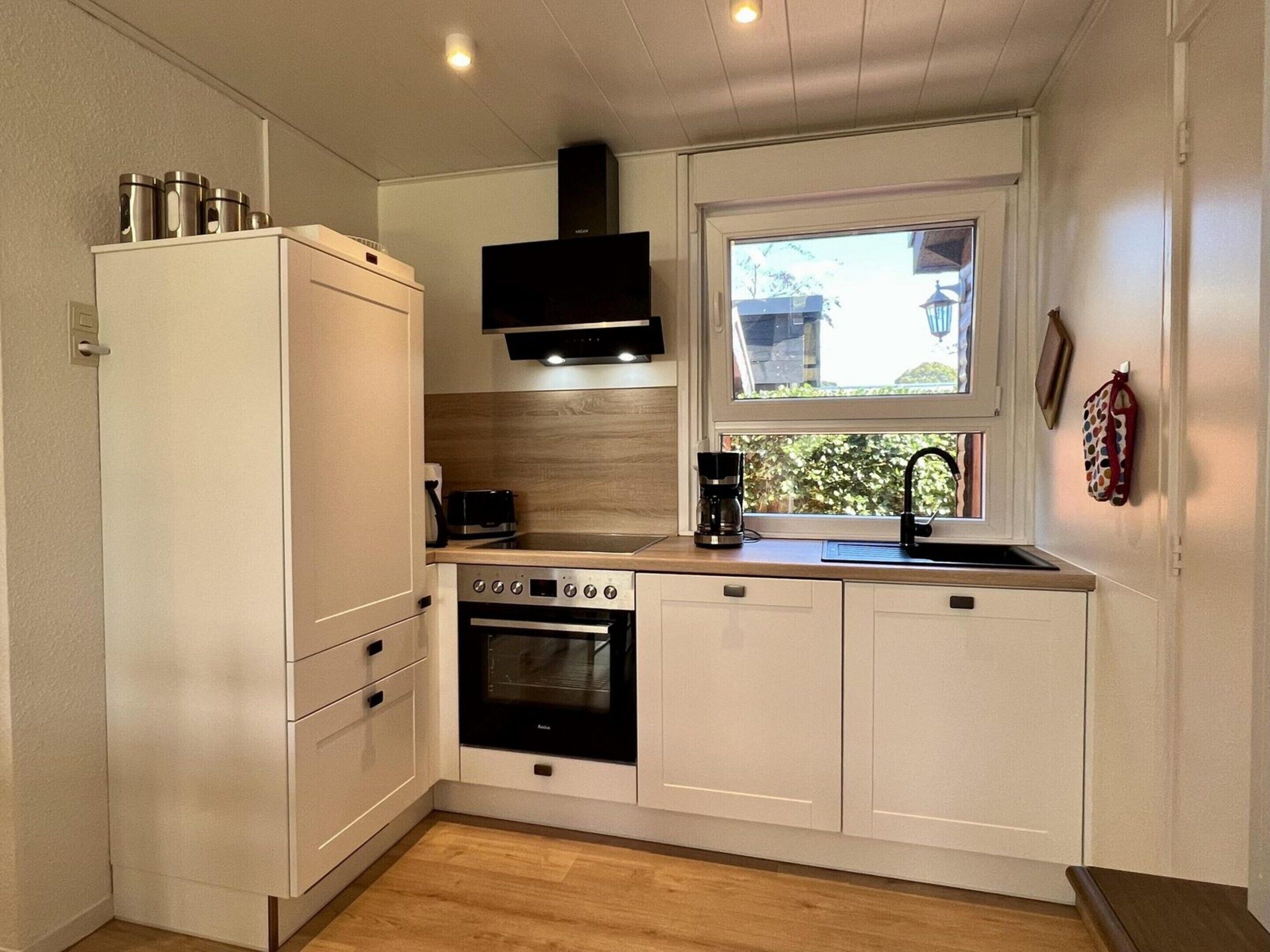 Una cocina compacta con gabinetes blancos, horno empotrado, cafetera y ventana con vista a zonas verdes.