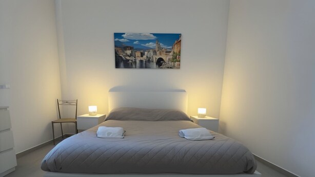 Une chambre moderne avec un grand lit, deux tables de chevet avec des lampes, un fauteuil et un tableau représentant un paysage urbain accroché au mur.