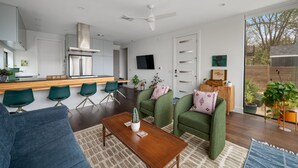 Une cuisine moderne dotée d’une cuisinière intégrée, un coin repas avec une table en bois et des chaises vertes. On y trouve également un ventilateur de plafond, un téléviseur à écran plat et une grande fenêtre donnant sur l’extérieur.