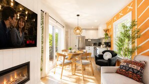 A modern living room with a fireplace, a dining area with a round table, and a kitchen with a stainless steel refrigerator.