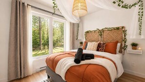 A bedroom with a large window, a bed with orange bedding, a hanging light fixture, and greenery on the walls.