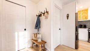 A hallway with white doors, a bench with a woven bag and a towel, a coat rack with a jacket, and a kitchen area with a refrigerator and stove.