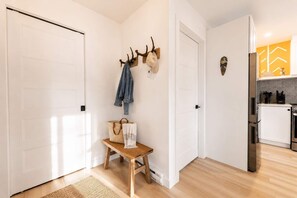 Un couloir avec des portes blanches, un banc avec un sac en osier et une serviette, un porte-manteau avec une veste, et un coin cuisine avec un réfrigérateur et une cuisinière.