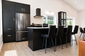 A modern kitchen with a stainless steel refrigerator, a central island with bar stools, and a black cabinet with glass doors. - Spa, Mountains & Ultimate Comfort | Le Jameson (Saint-Philémon)