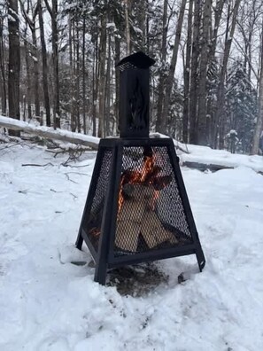 A snow-covered forest with a black metal fire pit in the center. - Spa, Mountains & Ultimate Comfort | Le Jameson (Saint-Philémon)