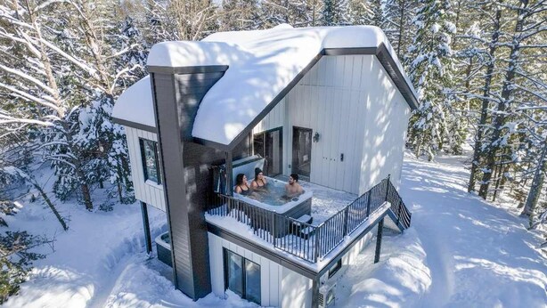 Eine moderne Holzhütte mit Whirlpool im zweiten Stock, umgeben von schneebedeckten Bäumen.