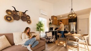 Ein Wohnzimmer mit einer Couch, ein Essbereich mit Stühlen, eine Küche mit einer Person beim Kochen und Wanddekoration.