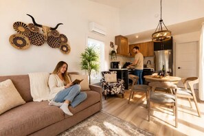 Een woonkamer met een bank, een eethoek met stoelen, een keuken waar iemand kookt en decoratieve wandkunst.