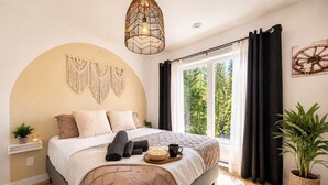 Ein Schlafzimmer mit einem Bett, einer Wandweberei aus Makramee, einer Hängelampe, einem Fenster mit Vorhängen und einer Pflanze.
