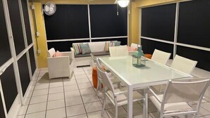 Een kamer met een glazen tafel, witte stoelen, een bank met kleurrijke kussens, een plafondventilator en een groot raam.