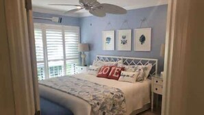 Een slaapkamer met een bed, een plafondventilator, een raam met jaloezieën en ingekaderde foto's aan de muur.
