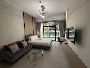 Quarto de hotel moderno com cama, sofá, televisão e janelas amplas.