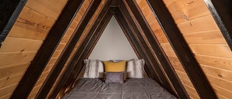 A triangular attic bedroom with a bed centered, wooden beams, and a sloped ceiling.