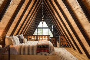 A cozy attic bedroom with a wooden sloped ceiling, a bed with a patterned blanket, and a window with a view.