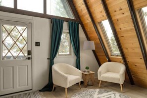 A cozy room with a sloped ceiling, a white door with a diamond-patterned window, two white armchairs, a small wooden side table, and a lamp.