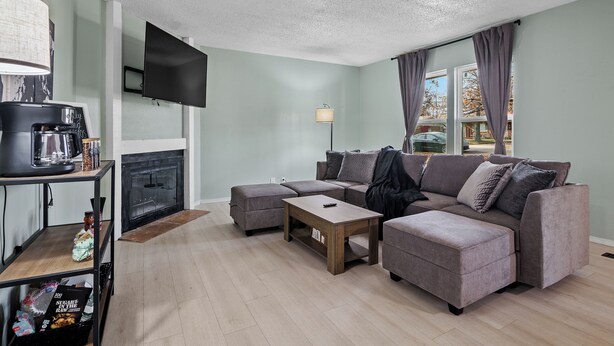 A living room with a sectional sofa, a coffee table, a fireplace, a TV mounted on the wall, and a window with curtains. - Charming Pastel Duplex in the Heart of Tulsa (Tulsa)