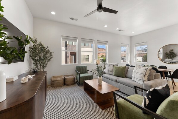 A modern living room with a sofa, armchairs, a coffee table, and a dining area. There are large windows providing natural light and views of the outside. - Villa #26 At Bloomington Country Club - Sunset Retreat 4 Bedroom Townhouse (St. George)
