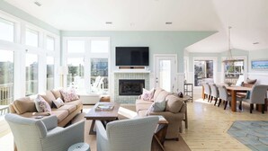 A spacious living room with a fireplace, large windows, and a dining area with a high table and chairs. - Newly Renovated! Semi Oceanfront w/ Pool & Hot Tub (Duck)