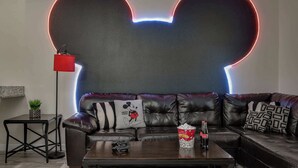Ein Wohnzimmer mit einer großen schwarzen Couch, einem Couchtisch mit einer Popcornschüssel und Flaschen, einer roten Stehlampe und einer Wand mit einem Neon-Mickey-Mouse-Motiv.