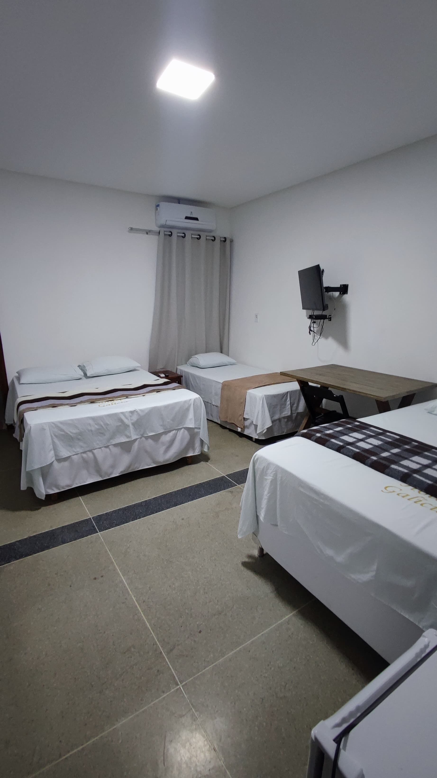 A hotel room with two single beds, a small table, a TV mounted on the wall, and an air conditioning unit.