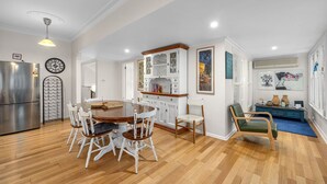 Un comedor con una mesa de madera y sillas blancas, una cocina con refrigerador y armarios, y una sala de estar con sofá y objetos decorativos.