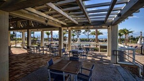 An outdoor patio area with tables and chairs, a pergola structure, and a view of palm trees and a body of water. - Oceanside Villa! Heated Pool, Hot Tub and Fitness Center (Hilton Head Island)