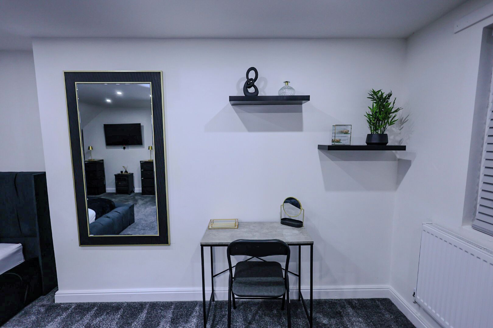 A compact room with a large mirror, a small table with a chair, and two wall-mounted shelves with decorative items.