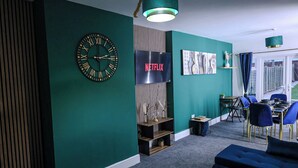 A modern living room with teal walls, a large clock, a Netflix sign, and a dining area with blue chairs. - Sleeps17 Bliss 7-bedroom Luxury Escape Birmingham (Birmingham)