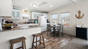 A kitchen with a central island, wooden stools, a dining table with chairs, and a view through the windows.