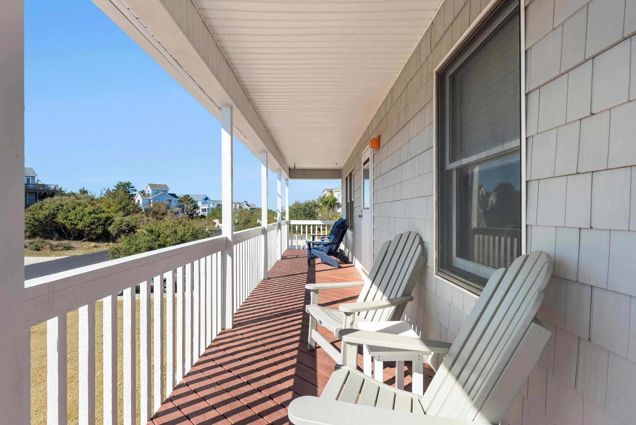 A covered porch with white railings, red wooden floor, and white Adirondack chairs.
