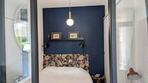 A bedroom with a bed, a headboard with a floral pattern, a wall-mounted shelf with framed pictures, a round light fixture, and a small table with a lamp. - Charming with Garden | Near train station (Rennes)