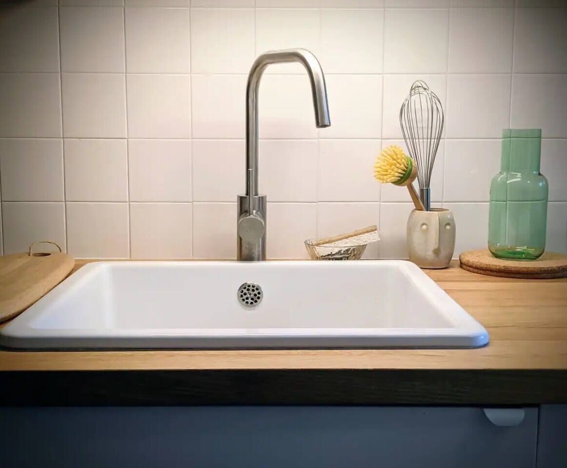 Une cuisine avec un évier blanc, un robinet en acier inoxydable, un plan de travail en bois, un fouet et une bouteille verte.