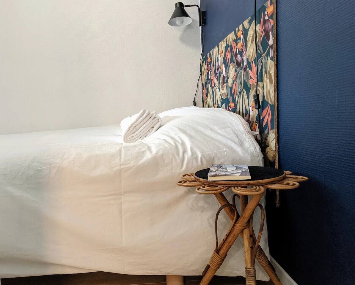 Un lit bien fait, recouvert d’une couette blanche et agrémenté d’un seul oreiller, une petite table d’appoint ronde en bois sur laquelle reposent un livre et une lampe, et une décoration murale florale aux couleurs vives.