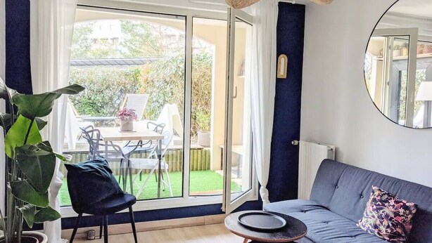 A modern living room with a sofa, a dining table, and chairs. There is a large window with curtains, a potted plant, and a hanging light fixture. - Charming with Garden | Near train station (Rennes)
