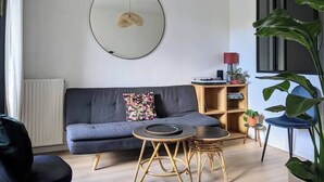 A modern living room with a grey sofa, a round mirror, a wooden coffee table, and a bookshelf with books and decorative items. - Charming with Garden | Near train station (Rennes)
