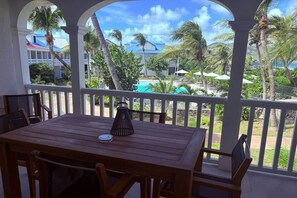 Mesa de madeira com cadeiras, uma luminária pequena e vista para uma piscina e palmeiras.