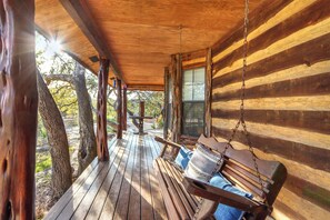 A wooden porch with a swing, a wooden table, and a view of trees and a building in the background. - Luxury Hill Country Escape • Pool, Hot Tub, Firepit • Near Wineries (Wimberley)