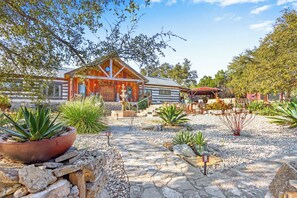 A rustic log cabin with a stone pathway leading to the entrance, surrounded by various desert plants and a small fountain.