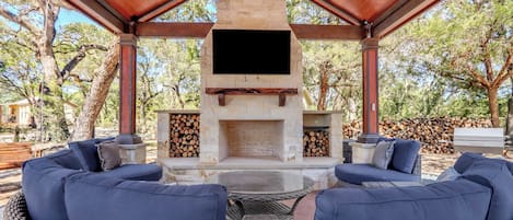 Un patio exterior con chimenea de piedra, asientos con cojines azules y un televisor de pantalla plana sobre el hogar.