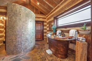 Un bagno con una colonna rivestita in mosaico, una vasca da bagno in rame, pareti in legno e una finestra che lascia entrare luce naturale.