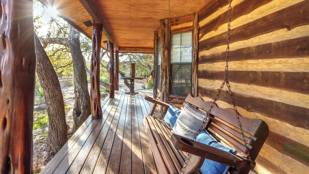 A wooden porch with a swing, a wooden table, and a view of trees and a building in the background. - Luxury Ranch w/ Pool, Hot Tub, Chef's Kitchen + Close to Wineries (Wimberley)