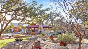 A rustic outdoor area with stone pathways, a wooden cart, and a rustic building with a red roof. - Luxury Ranch w/ Pool, Hot Tub, Chef's Kitchen + Close to Wineries (Wimberley)