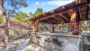 An outdoor bar area with stone walls, metal bar stools, and a wooden pergola with string lights. - Luxury Ranch w/ Pool, Hot Tub, Chef's Kitchen + Close to Wineries (Wimberley)