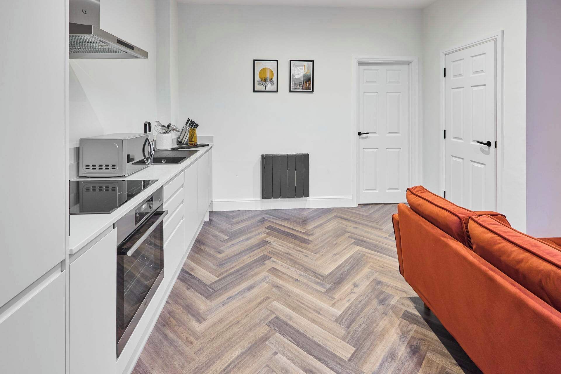 A modern kitchen with white cabinetry, a microwave, and a stove. There is a herringbone-patterned wooden floor and a red sofa in the living area.