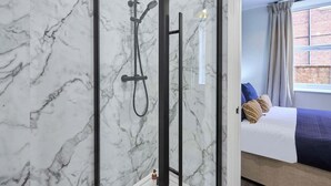 Ein Badezimmer mit Dusche, Marmorwänden und ein angrenzender Raum mit einem Bett und einem blau-braunen Kopfteil.
