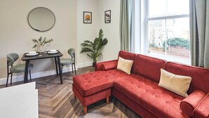 A living room with a red sectional sofa, a dining table with chairs, a potted plant, and a large window with curtains. - Host & Stay | Apartment 3, Upton House (Northampton)