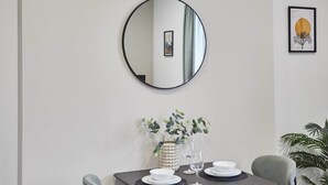 A dining area with a round mirror on the wall, a framed picture, a table set with plates and a vase of greenery, and a potted plant. - Host & Stay | Apartment 3, Upton House (Northampton)